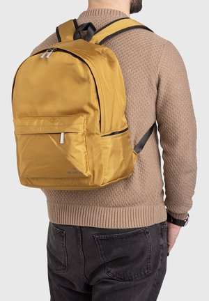 Mann mit braunem Pullover und schwarzer Jeans, der einen senffarbenen Rucksack mit Vordertasche und schwarzen verstellbaren Gurten trägt.