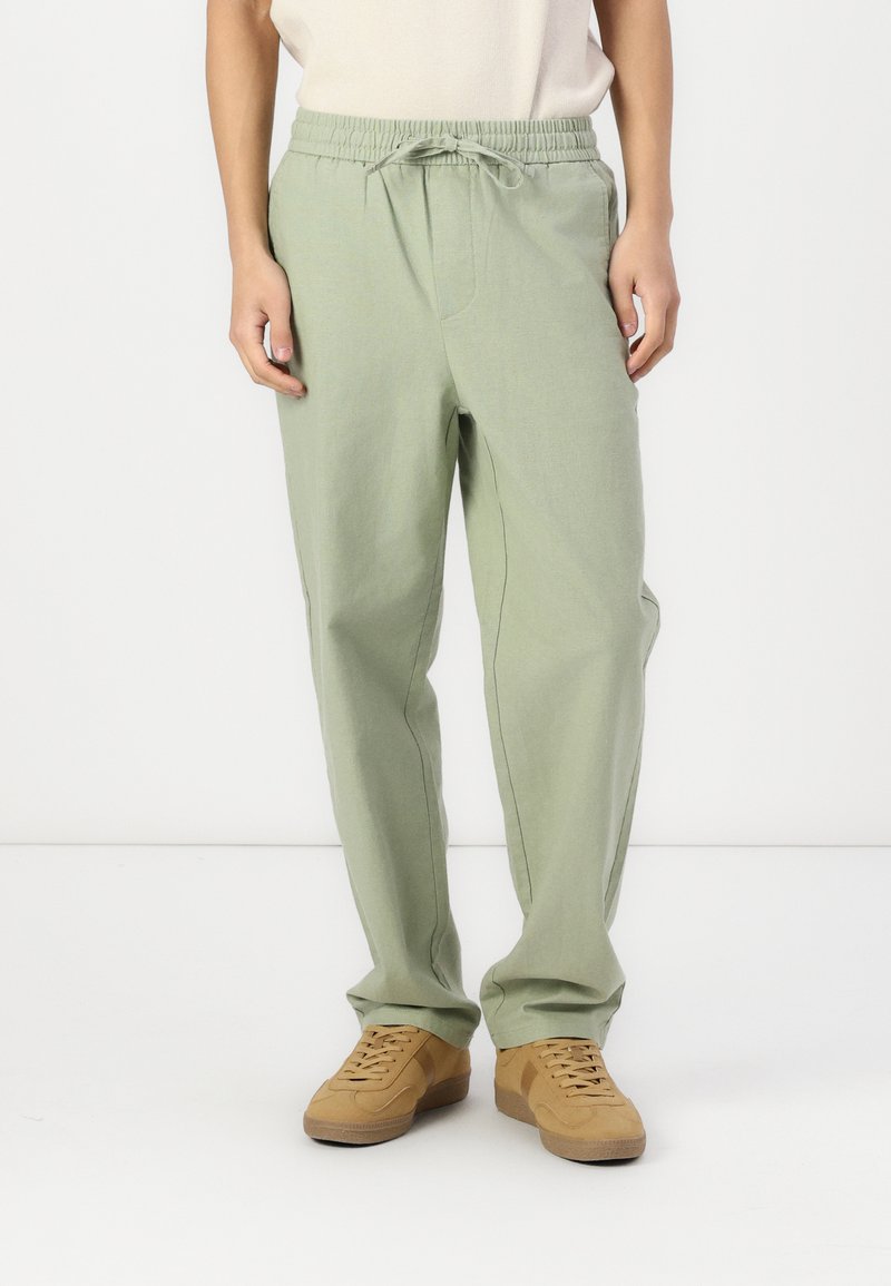Pantalones sueltos de color verde claro con cintura elástica y cordón. Textura suave con bolsillos laterales. Combinados con zapatillas de deporte color beige.