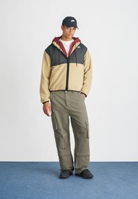 Κρέμ και μαύρο φερμουάρ μπουφάν με κουκούλα και μαρουνί λεπτομέρειες, σε συνδυασμό με πράσινα cargo παντελόνια με πολλές τσέπες και μαύρα παπούτσια.