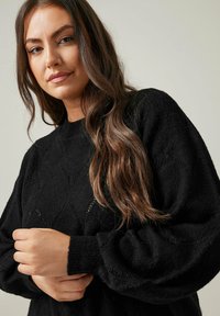 Maglione nero a maglia con motivo a losanga, maniche lunghe a sbuffo e polsini a costine. La texture appare morbida e leggera.
