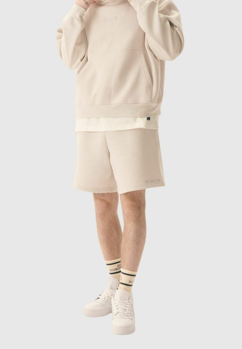 Haut à capuche beige avec une poche frontale, associé à un short assorti. Baskets blanches et chaussettes avec une bande noire. Texture douce, design décontracté.