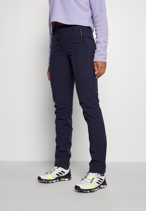Mørkeblå bukser med slim fit, der har to lynlås-lommer. Modellen sætter dem sammen med hvide sneakers med sorte og neongule accenter.