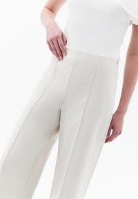 Béžové kalhoty s širokými nohavicemi a prošívanými detaily, vysokým pasem a bočními kapsami, doplněné přiléhavým bílým topem. Hladká textilie.