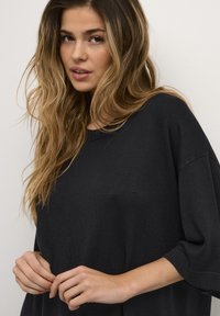 Maglione nero oversize con maniche corte, scollatura rotonda e tessuto lavorato a maglia. I capelli sono lunghi e ondulati, incorniciando il viso.