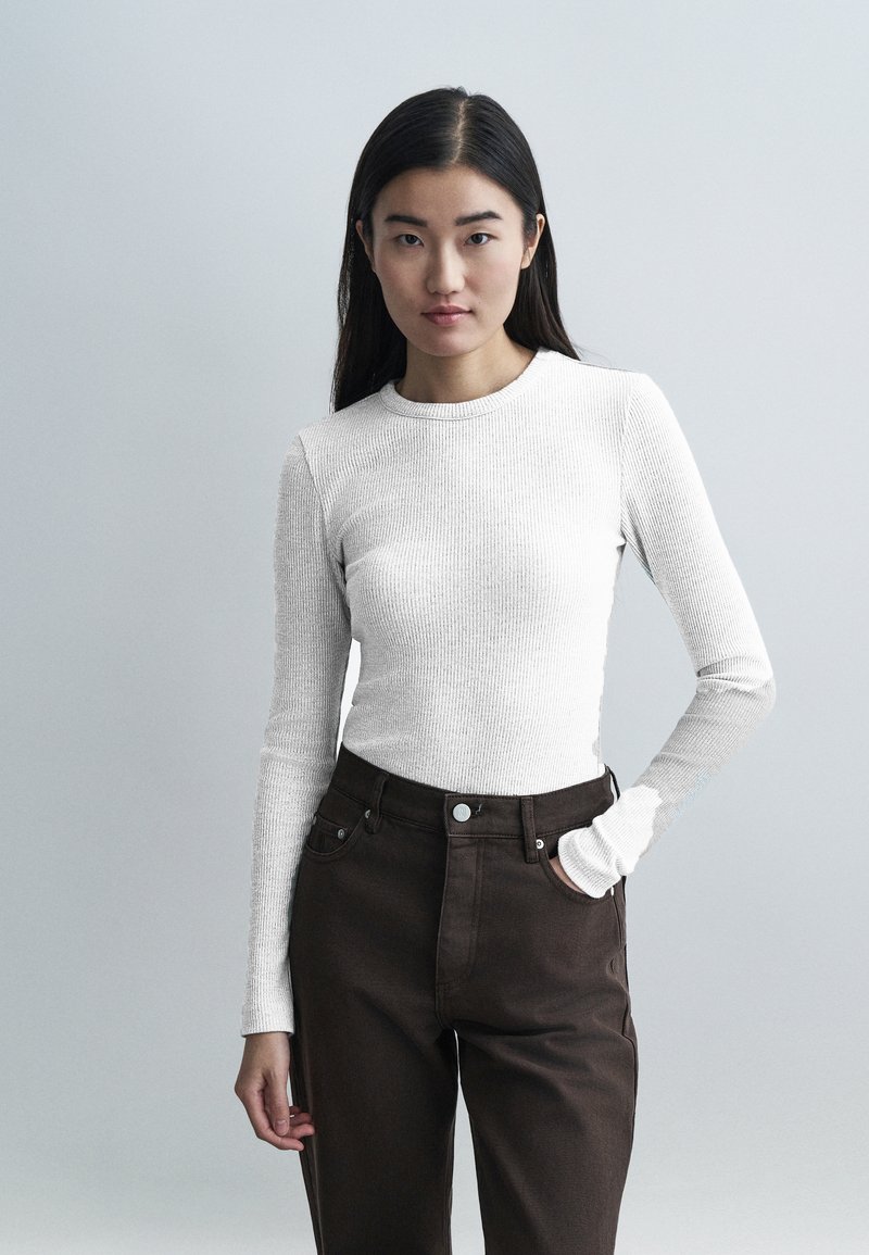 Valge soonikkoes pika varrukaga top ümmarguse kaelusega, kombineeritud kõrge vöökohaga tumedate pruunide teksadega. Lihtne disain ja kitsas lõige.