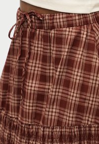 Κόκκινη και μπεζ καρό φούστα με υφασμάτινη υφή, ελαστική μέση και κορδόνι στη λεπτομέρεια. Το βολάν στο τελείωμα προσθέτει όγκο και σχήμα.