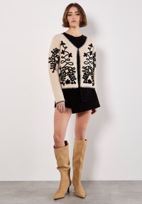 Beiger Cardigan mit schwarzer floralem Stickerei, V-Ausschnitt und kontrastierenden Besätzen. Kombiniert mit einem schwarzen Kleid und beigen Stiefeln bis zu den Knien.