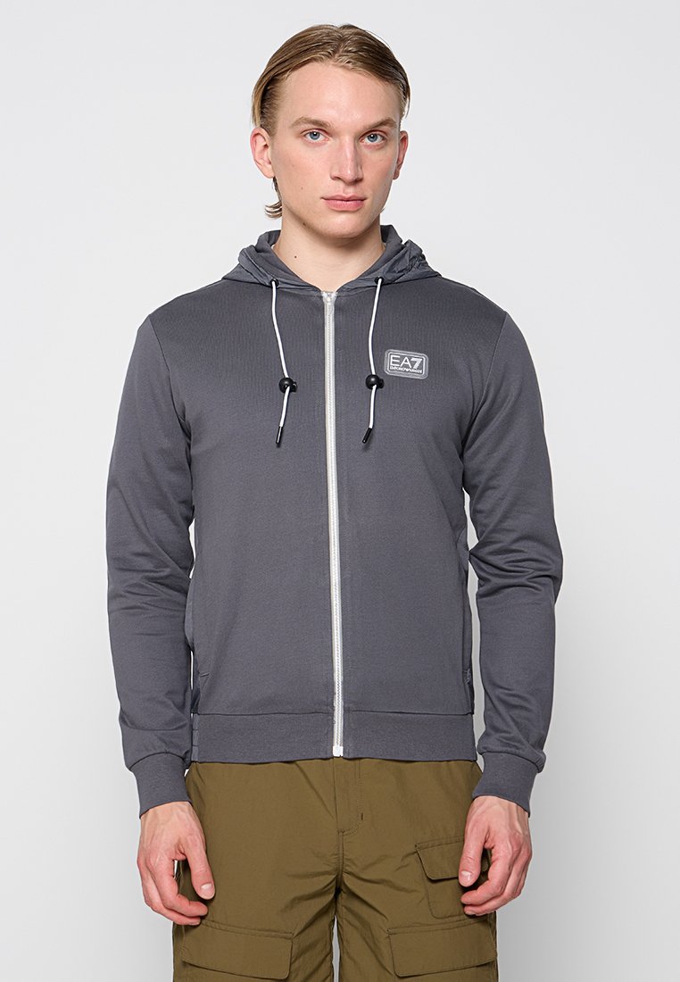 Giovane uomo che indossa una felpa grigia con zip e coulisse bianche e pantaloni cargo color oliva, in piedi contro uno sfondo chiaro e neutro.