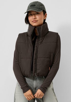 Junge Person trägt eine dunkle Steppweste über einem Langarmshirt, hell gewaschene Jeans und eine dunkle Baseballkappe, steht mit verschränkten Händen.