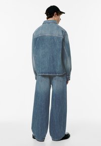 Lichtblauwe denimjack met een relaxte pasvorm en lange mouwen, gecombineerd met bijpassende oversized denimbroek. Zwarte pet en stevige zwarte laarzen.