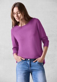 Fioletowy sweter z długim rękawem i okrągłym dekoltem, z prążkowanymi mankietami, połączony z jasnoniebieskimi dżinsami z przednimi kieszeniami i wyblakłym wykończeniem.