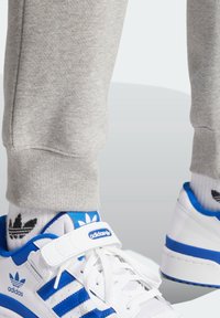 Vita sneakers med blå accenter och tre ränder, åtföljda av grå träningsbyxor. Strumpor med ett svart- och vitt mönster av löv synliga.