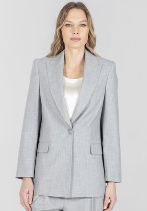 Kobieta w jasnoszarej, dopasowanej marynarce z wyciętymi klapami i zapięciem na guziki z przodu, ubrana na biało, na jednolitym tle.