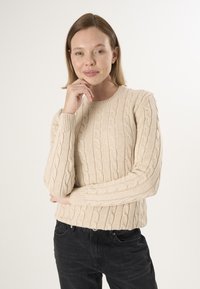 Beige Kabelstrickpullover mit Rundhalsausschnitt und gerippten Bündchen, getragen mit schwarzen Jeans, die über strukturierte Muster und einen lässigen Schnitt verfügen.