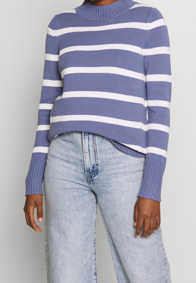 Lila und weiße gestreifte Strickjacke mit hohem Kragen und gerippten Bündchen, getragen mit hellblauen, weiten Jeans. Flache Textur.