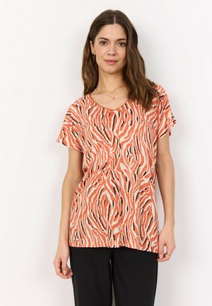 Kvinna med vågigt brunt hår som bär en korallfärgad, beige och svart zebra-randig kortärmad topp tillsammans med svarta byxor, stående mot en vit bakgrund.