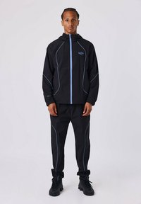 Chándal deportivo negro que incluye una chaqueta con cremallera y pantalones con ribetes azul claro. La chaqueta tiene capucha y puños elásticos. Tejido texturizado.