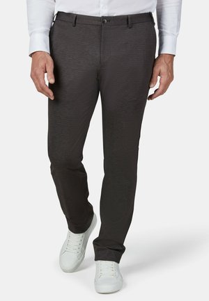 Mann trägt dunkelgraue, strukturierte Anzugshosen, ein weißes Hemd und weiße Turnschuhe und steht vor einem schlichten Hintergrund.