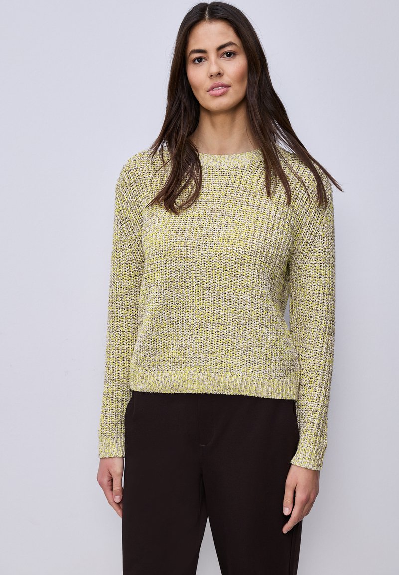 Femme aux longs cheveux bruns portant un pull en maille jaune et gris et un pantalon noir, debout devant un fond uni gris clair.