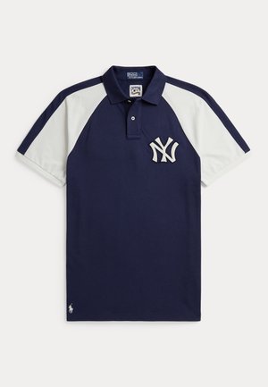 Mężczyzna i kobieta stoją obok siebie, ubrane w granatowe i białe koszulki polo New York Yankees oraz szare spodnie dresowe z rękami w kieszeniach.
