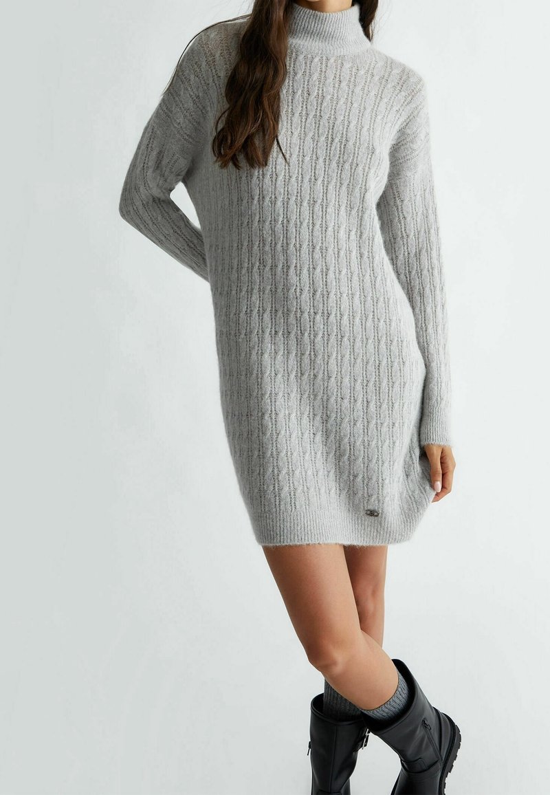 Femme portant une robe pull à col roulé en maille torsadée gris clair avec des chaussettes grises et des bottes noires mi-mollet, debout devant un fond uni.