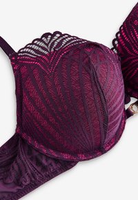 Fialová krajková podprsenka se zvlněnými okraji a složitými geometrickými vzory na košíčcích a pásku, ukazující detailní texturu a síťovanou látku.