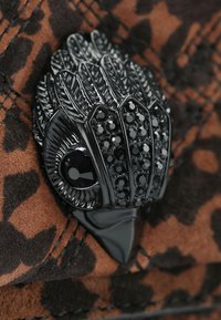 Spilla a forma di uccello in smalto nero con dettagli in piuma e embellimenti in pietra nera, poggiata su uno sfondo a stampa leopardata marrone.