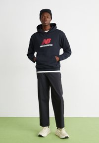 Tengerészeti kapucnis pulóver piros New Balance logóval, fekete nadrág és bézs sneaker. Az öltözék laza dizájnnal és kényelmes fit-tel rendelkezik.