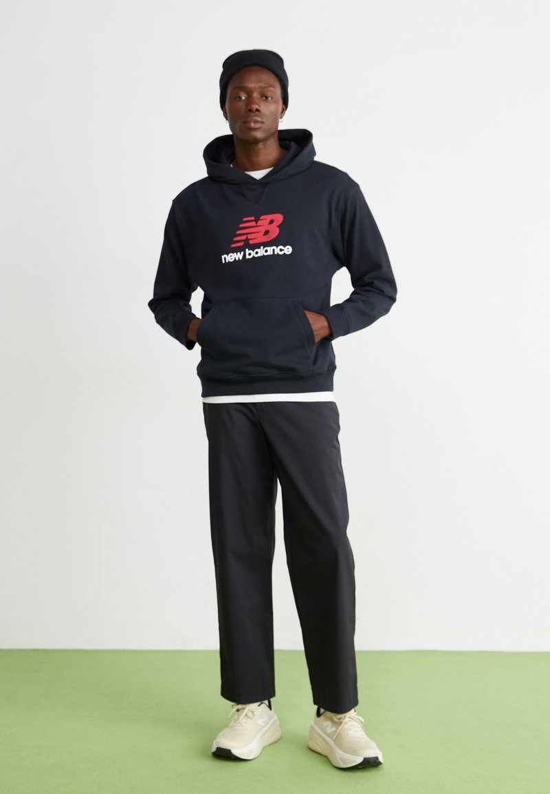 Marineblauer Hoodie mit rotem New Balance-Logo, schwarze Hose und beigen Sneakern. Das Outfit hat ein lässiges Design und einen entspannten Schnitt.
