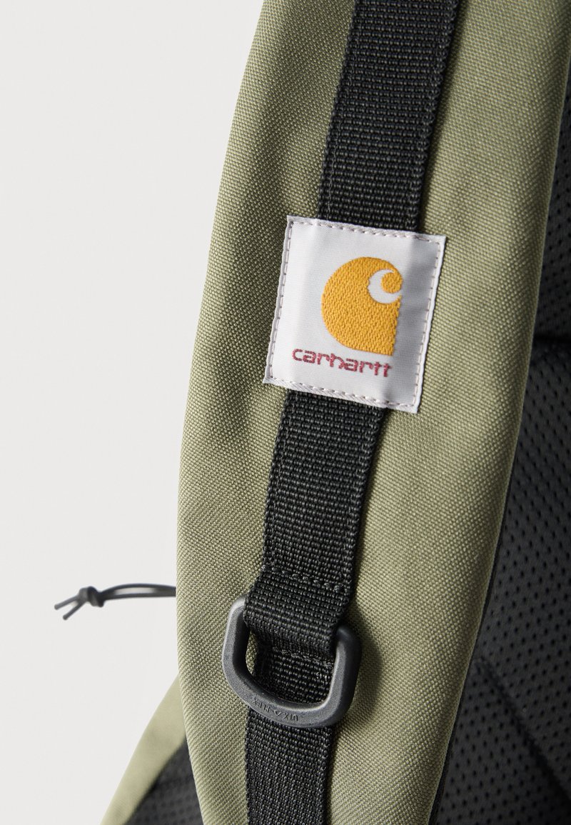Grüner Rucksackriemen mit schwarzer Befestigungsschlaufe und weißem Carhartt-Logo-Patch mit gelbem Emblem und rotem Text.