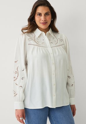 Kvinde iført hvid skjorte med knapper og broderede udskæringsdetaljer på ærmer og bryst, kombineret med blå jeans, stående foran en ensfarvet væg.