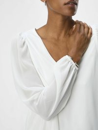 Blouse blanche avec un décolleté en V profond et des manches bouffantes en voile. Tissu lisse avec une texture douce. Accompagnée d'un simple bracelet en argent.