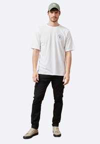 Camiseta blanca de algodón con mangas cortas y gráfico azul, combinada con pantalones cargo negros y zapatillas beige. Gorra verde a juego.