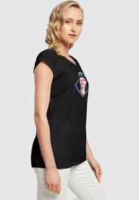 Schwarzes T-Shirt mit kurzen Ärmeln, das ein gesticktes Frida Kahlo Design in lebendigen Farben auf einem rhombusförmigen Patch zeigt.