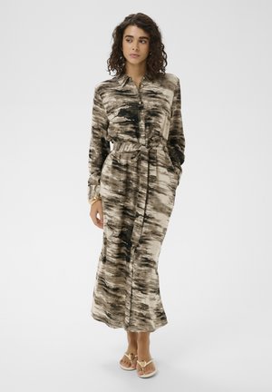 Femme aux cheveux bouclés portant une longue robe marron et beige à motifs, boutonnée, avec une ceinture à la taille et des sandales beiges, debout devant un fond uni.