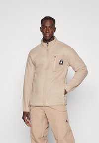 Beige fleecejakke med høy krage, hel glidelås og brystlomme. Har et logo-patch og teksturert stoff. Kombinert med beige bukser.