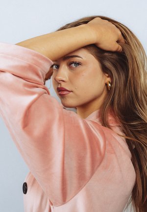 Noor naine, kellel on pikad juuksed ja kes kannab roosat sametist jakki, tõstab ühe käe pea kohale ja vaatab küljele neutraalse ilmega.