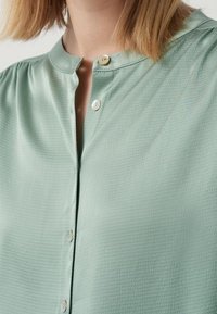 Blusa verde menta con scollo rotondo, chiusura a bottoni e tessuto testurizzato con leggere ondulazioni. Gli accenti includono bottoni bianchi.