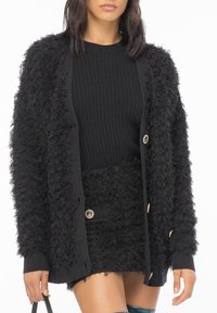 Schwarze, flauschige Strickjacke mit gerippten Bündchen, ausgestattet mit goldenen Knopfakzenten. Getragen über einem schwarzen, gerippten Oberteil und einem passenden, strukturierten Rock.