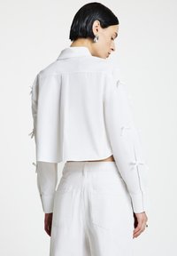 Frau mit kurzen schwarzen Haaren, die ein weißes, bauchfreies Shirt mit Schleifen an den Ärmeln und eine hochtaillierte weiße Hose trägt, seitlich vor weißem Hintergrund stehend.