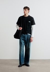 Νεαρός άνδρας που στέκεται, φορά πολλαπλά μαύρα και λευκά μακρυμάνικα πουκάμισα, άνετα μπλε τζιν, μαύρα παπούτσια και κρατά μια μαύρη τσάντα ώμου.