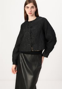 Black textured jacket featuring a round neckline, oversized sleeves, and decorative toggle closures, paired with a sleek black leather skirt.