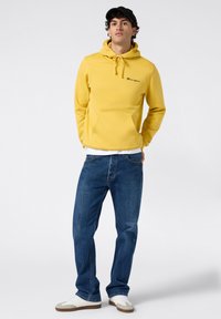 Felpa gialla con tasca frontale e cordoncino, logo sul petto. Abbinata a jeans blu e sneakers con dettagli beige. Sfondo semplice.