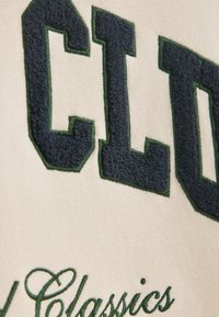 Un tessuto color crema presenta lettere in rilievo di colore blu navy contornate di verde scuro, con ulteriori dettagli ricamati in verde sottostanti.