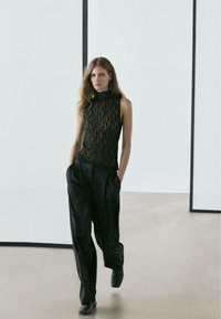 Zwart kanten mouwloze top met een hoge halslijn, gecombineerd met losse zwarte broek en zwarte schoenen, tegen een minimalistische achtergrond.