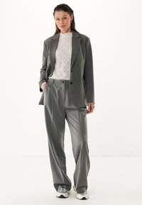 Traje gris a medida que presenta un blazer de un solo botón y pantalones sueltos. Debajo, una blusa de encaje blanca con cuello alto. Zapatillas plateadas complementan el look.