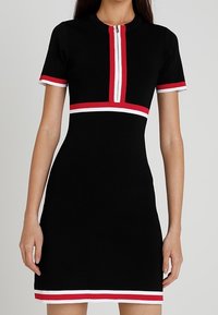 Femme portant une robe noire à manches courtes avec des bordures rouges et blanches, dotée d'une fermeture éclair à l'avant et d'une taille cintrée.