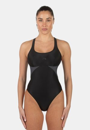 Donna in piedi, rivolta in avanti, indossa un costume intero nero con pannelli laterali grigi, capelli legati, espressione neutra, sfondo semplice.