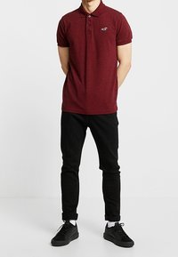 Mörkröd pikétröja med korta ärmar och krage, med en liten logotyp. Tillsammans med svarta slim-fit byxor och svarta sneakers.