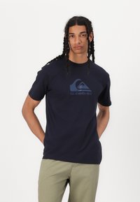 Tums zilnā krāsa, izgatavots no kokvilnas, ar drukātu viļņu logotipu un vārdu "Quiksilver" kontrastējošā zilā krāsā. Īsas piedurknes un apaļa kakla izgriezums.